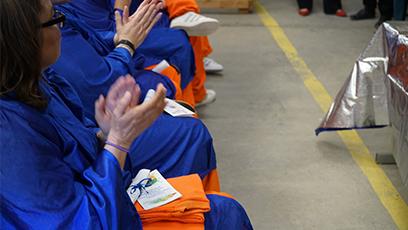 A group of individuals in blue graduation gowns and orange pants are seated in a row, clapping. The focus is on their hands and laps, with one person holding a colorful program booklet and folded orange fabric. The scene is part of a graduation ceremony taking place at Arizona State Prison Complex-Perryville