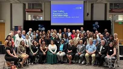 A large group of people pose together for a photo during the East Valley Rotaract Club charter celebration at Rio Salado College. The attendees, dressed in business and semi-formal attire, fill the stage area and front rows of the event space. Behind them, a projection screen displays the event title and date: “East Valley Rotaract Club at Rio Salado College, Charter Celebration, Thursday, July 25, 2025.” An American flag and balloon decorations are also visible in the background.