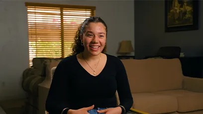 Alexandria Woodruff sits smiling on a couch in a living room, wearing a black long-sleeve top, speaking confidently during an interview for Rio Salado College’s spring campaign.