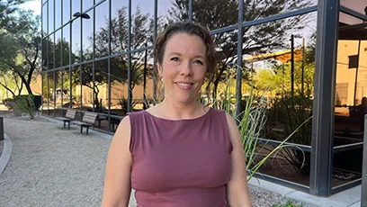 Alysa Call, with light brown hair pulled back stands outdoors in front of a reflective glass building. She is wearing a sleeveless mauve top and smiling at the camera, with desert landscaping and trees in the background.