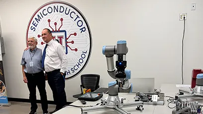 Two men stand together smiling in a classroom lab with a large ‘Semiconductor High School’ logo on the wall behind them. In the foreground, a robotic arm and electronic equipment sit on a worktable, highlighting the technology-focused environment.