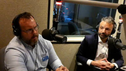 Ken Wilk and Zach Mannheimer wearing headphones speak into microphones during a podcast recording in a radio studio.