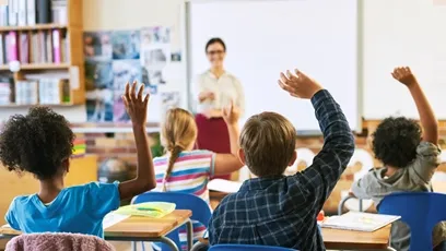 Students raise their hands to participate in a classroom discussion while a teacher stands at the front of the room.