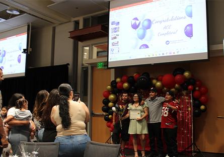 A female graduate poses with four men, likely family members, in front of a festive balloon arch featuring red, black, and gold balloons. She holds a certificate while the others smile with arms around each other. Several guests gather to the side, with one woman holding up a phone to take a photo and another cradling a child. A slide projected above reads “Congratulations, ACE Completers!” with the Rio Salado College logo, affirming the celebratory context of the event.