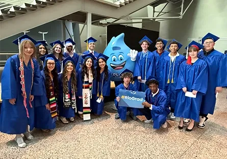 A group of Rio Salado College dual enrollment students, dressed in blue graduation gowns and caps, pose together before the commencement ceremony. They are gathered around Splash, the Rio Waves mascot, a large blue cartoon water droplet giving a friendly wave. Two students in front hold a sign that reads “#RioGrad.” The group stands indoors near a staircase, smiling and excited for the event.