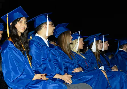 A row of graduates in royal blue caps and gowns sit side by side during a commencement ceremony. Most are looking forward, while one young woman near the front smiles at the camera. The lighting highlights the graduates against a dark background, emphasizing the celebratory moment.