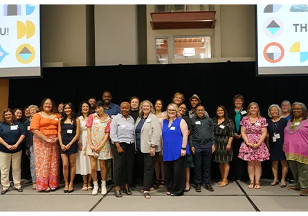 A large group of people poses on a stage at the Educator Preparation Program convocation. The diverse group includes students, faculty, and staff, smiling in front of a black curtain. Two large screens on either side display colorful graphics and the words “THANK YOU!” The atmosphere is celebratory and supportive, recognizing the accomplishments of future educators.