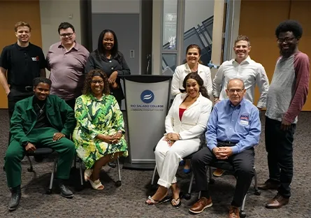 Eleven attendees of the small business networking event pose together for a group photo in front of a Rio Salado College podium. They are dressed in a mix of business and casual attire, smiling warmly at the camera in a spacious event room with wood-paneled doors in the background.