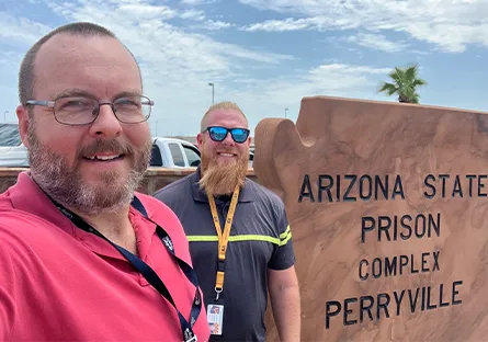 A selfie of two smiling professionals standing outdoors in front of a sandstone sign reading “Arizona State Prison Complex – Perryville.” On the left is Ken Wilk, faculty chair for applied technology and engineering, wearing glasses, a short-cropped beard, and a red polo shirt. On the right is Casey Werhanowicz, Program Director at ASPC‑Lewis, with a trimmed reddish-gold beard, sunglasses, and a dark shirt with a yellow‑striped lanyard. A palm tree and blue‑sky background appear behind them.