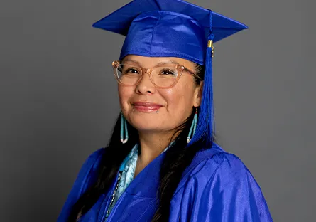 A graduate wearing a blue cap and gown smiles softly while posing against a gray background. She is wearing glasses, turquoise earrings, and a matching necklace, giving a proud and celebratory appearance.