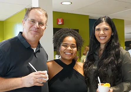 Three Rio Salado College employees smile together while holding cups of food at an indoor gathering. The man on the left wears glasses and a black polo shirt, the woman in the center has curly hair and a black top, and the woman on the right has long dark hair and a green blouse. They appear relaxed and cheerful in a brightly lit office space with lime green walls in the background.