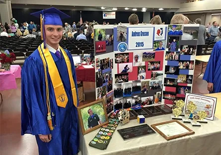 A young graduate wearing a blue cap and gown with a gold stole smiles while standing next to a display board at a graduation event. The board, labeled “Andreas Grossen 2015,” is decorated with numerous photos, certificates, and awards highlighting his achievements. Trophies, plaques, and personal memorabilia are arranged on the table in front of the display. Other graduates and families can be seen seated and mingling in the background of the large hall.