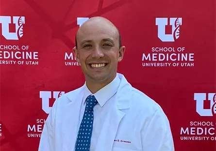 A man wearing a white medical coat and blue tie smiles in front of a red backdrop featuring the University of Utah logo and the words “School of Medicine, University of Utah.”