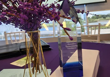 A crystal award engraved with “Institutional Award for Entrepreneuring Excellence – 2025 – Rio Salado College” sits on a table draped in purple. Next to it is a decorative glass vase with gold geometric trim, filled with branches of small purple blossoms. In the background, a projection screen and large windows with views of greenery and outdoor structures are visible, creating a celebratory event setting.
