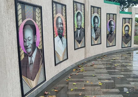 A curved outdoor wall displays framed portraits of historical figures in colorful, geometric designs. The walkway in front of the wall is wet with scattered leaves, suggesting recent rain.