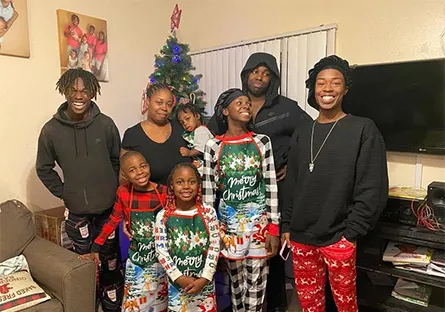 A family stands together smiling in a living room decorated for Christmas, with a small Christmas tree in the background. Several children wear festive “Merry Christmas” pajamas while the adults stand behind them in casual clothing. The joyful group poses for a holiday photo, capturing a warm family moment.