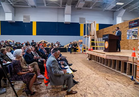 A speaker stands at a podium addressing an audience seated in a construction training facility. The room features wood framing displays, safety cones, and banners for NCCER accreditation and workforce development partners.