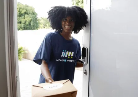 Smiling Meals on Wheels volunteer wearing a navy T-shirt delivers a boxed meal while standing at an open front door.