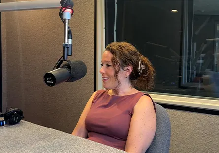 Alysa Call sits at a table in a recording studio, speaking into a large studio microphone. She is wearing a sleeveless mauve top and smiling while looking toward the right.