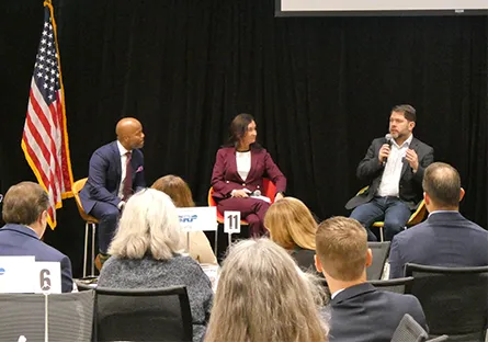 Three panelists sit on stage during a discussion in front of an audience. From left to right: Tempe Mayor Corey Woods, Tempe Chamber President & CEO Robin Arredondo-Savage, and Arizona Senator Ruben Gallego. Gallego is speaking into a microphone while the others listen. An American flag stands to the left, and attendees are seated at tables in the foreground.