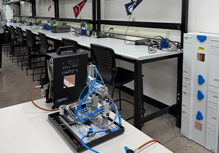 Semiconductor lab classroom with rows of workstations equipped with tools and electronic devices. In the foreground, a compact training system with blue tubing and mechanical components sits on a table, while lockers and additional lab benches line the back wall.