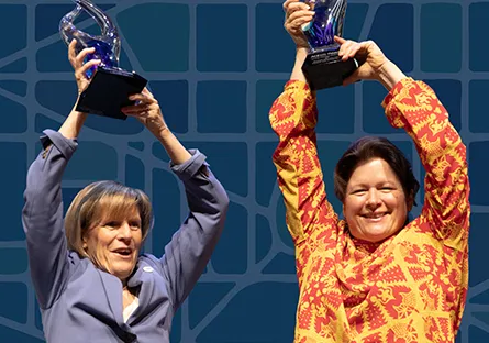 Two women smiling and raising glass awards above their heads against a blue patterned background.