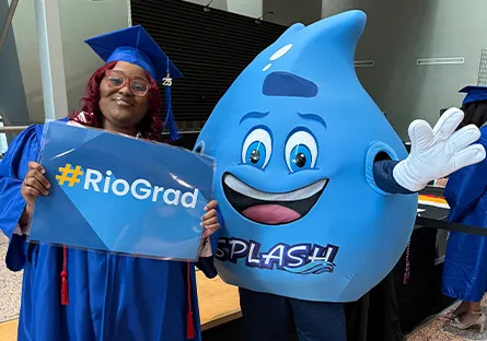 Graduate Kevina Aubrey in a blue cap and gown holding a “#RioGrad” sign stands next to Splash, the Rio Salado College mascot, who is waving.