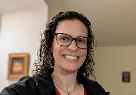 A woman with curly hair and glasses smiles at the camera in a home setting, wearing a black jacket over a red striped top.