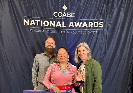 Three people stand smiling in front of a backdrop reading “COABE National Awards Honoring Excellence in Adult Education,” with one person holding an award.
