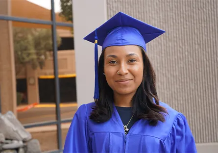 Portrait of Ebony Livas wearing a blue graduation cap and gown, standing outdoors and looking at the camera.