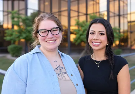 Maren “Rain” Colon, left, and Ariel Davila, right, smile together outdoors in front of a glass building and greenery.