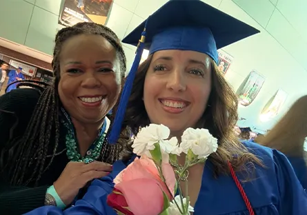 Rio Salado College graduate Diana Loya, wearing a blue cap and gown, smiles beside Roxie, who has her arm around Diana's shoulder. Diana holds a bouquet of pink and white flowers at the graduation celebration.