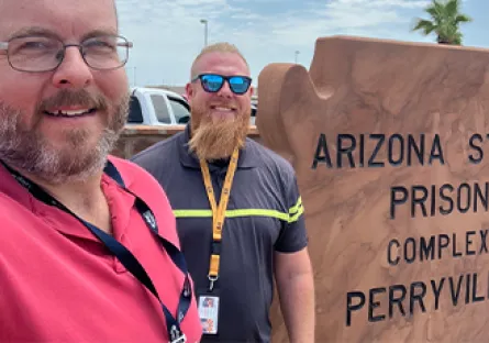 A selfie of two smiling professionals standing outdoors in front of a sandstone sign reading “Arizona State Prison Complex – Perryville.” On the left is Ken Wilk, faculty chair for applied technology and engineering, wearing glasses, a short-cropped beard, and a red polo shirt. On the right is Casey Werhanowicz, Program Director at ASPC‑Lewis, with a trimmed reddish-gold beard, sunglasses, and a dark shirt with a yellow‑striped lanyard. A palm tree and blue‑sky background appear behind them.