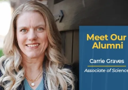 Carrie Graves, a blonde woman with long wavy hair, smiles while standing outdoors next to a dark wooden wall. She is wearing a navy blazer over a light blue top and layered necklaces. A blue graphic box on the right reads: ‘Meet Our Alumni — Carrie Graves, Associate of Science.’