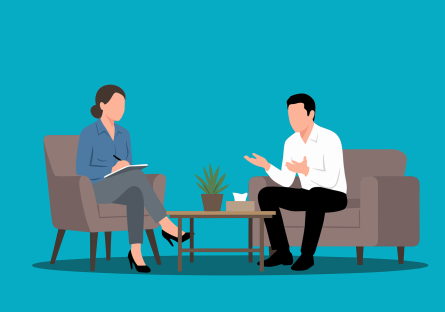 A man speaking to a woman therapist; both of them are seated with a table between them with a potted plant on it. Behind them is a solid aquamarine blue background