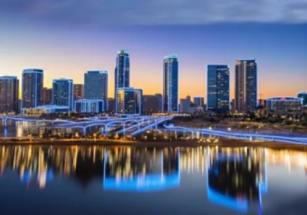 Downtown skyline at dusk with illuminated high-rise buildings and their reflections shimmering in the water along a riverfront.